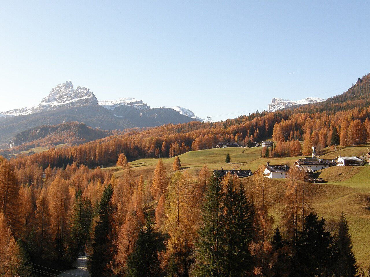 Boschi di larici color rame e prati ancora verdi intorno ad alcune case, con cime innevate sullo sfondo, a Cortina d'Ampezzo in autunno