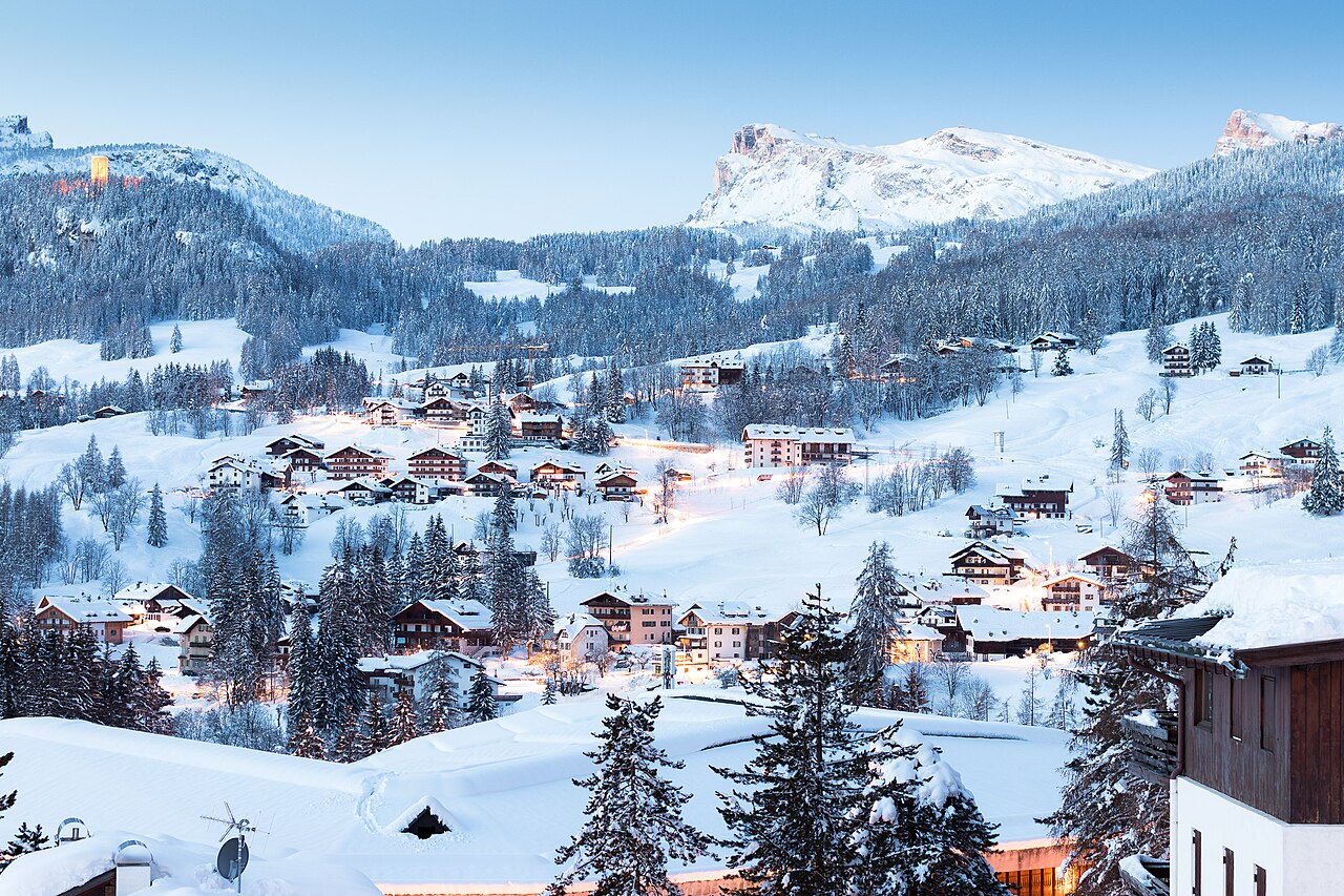 Case e boschi di Cortina d'Ampezzo coperti di neve alle prime luci del giorno, con i lampioni ancora accesi e le cime innevate sullo sfondo