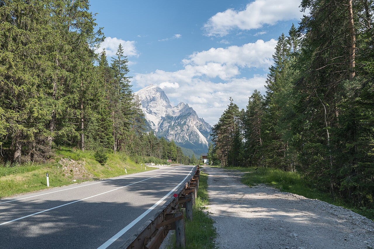 La statale 51 di Alemagna tra i boschi a sud di Cortina d'Ampezzo, con la ciclabile sterrata sulla destra e una cima dolomitica sullo sfondo